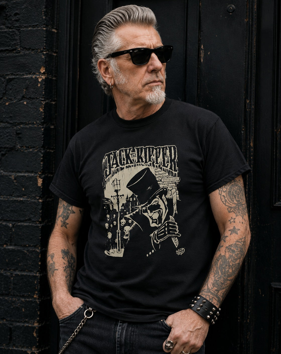 A man with gray hair, sunglasses, and tattoos leans against a black wooden door and brick wall, wearing a black graphic t-shirt with a beige 'Jack the Ripper' illustration.