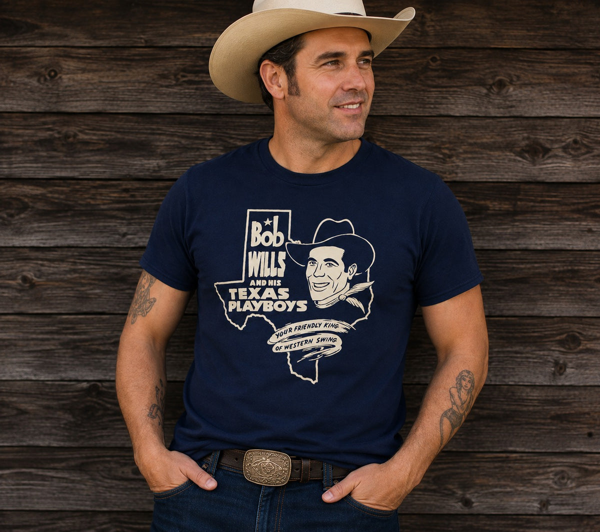 A man wearing a navy blue graphic t-shirt with a white print of Bob Wills and the Texas Playboys, paired with a beige cowboy hat and jeans, standing against a rustic dark wood plank wall.