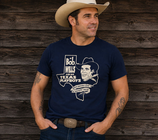 A man wearing a navy blue graphic t-shirt with a white print of Bob Wills and the Texas Playboys, paired with a beige cowboy hat and jeans, standing against a rustic dark wood plank wall.