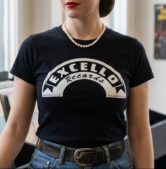 A woman with a 1950s-style hairstyle stands in a record store wearing a black graphic t-shirt that reads 'EXCELLO RECORDS'. The background features vintage posters of Elvis Presley and Wanda Jackson, and shelves of vinyl records.