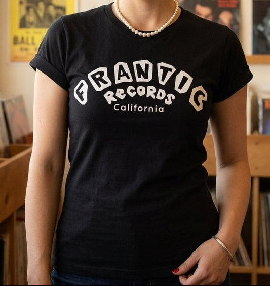 A woman with vintage-style dark hair stands in a record store wearing a black graphic t-shirt that reads 'FRANTIC RECORDS California' in white. The background features wooden shelves filled with vinyl records and posters.