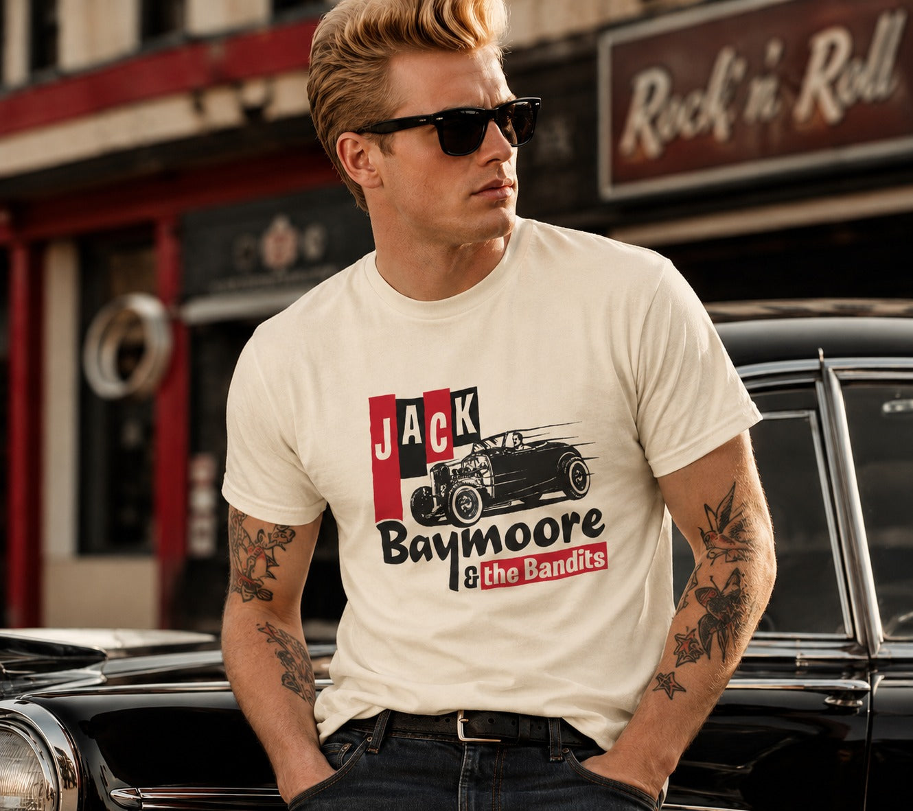 A man wearing a cream-colored graphic t-shirt with a 'Jack Baymoore & the Bandits' hot rod car design, standing in front of a vintage black car in a street setting.