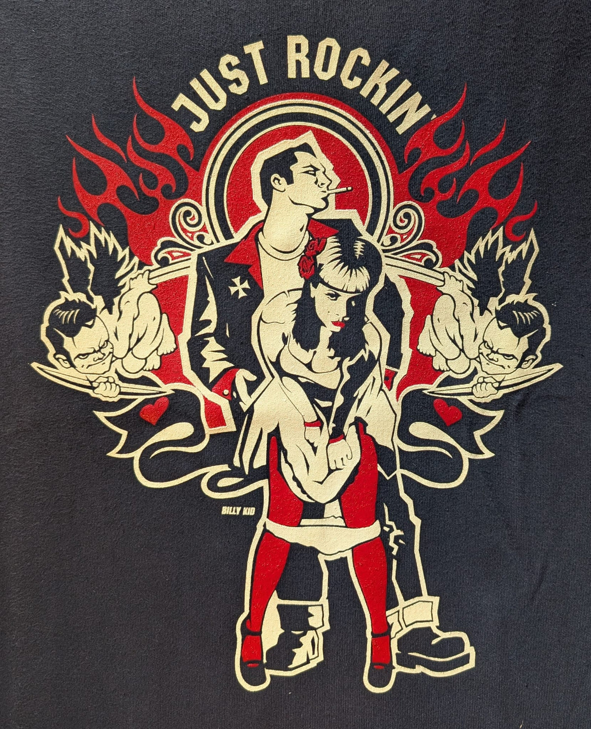 A close-up of a graphic t-shirt featuring a retro punk-style illustration with the text 'JUST ROCKIN' at the top. The design shows a central couple with a cigarette, flanked by two muscular figures holding microphones, all in black, red, and cream, on a dark charcoal background.
