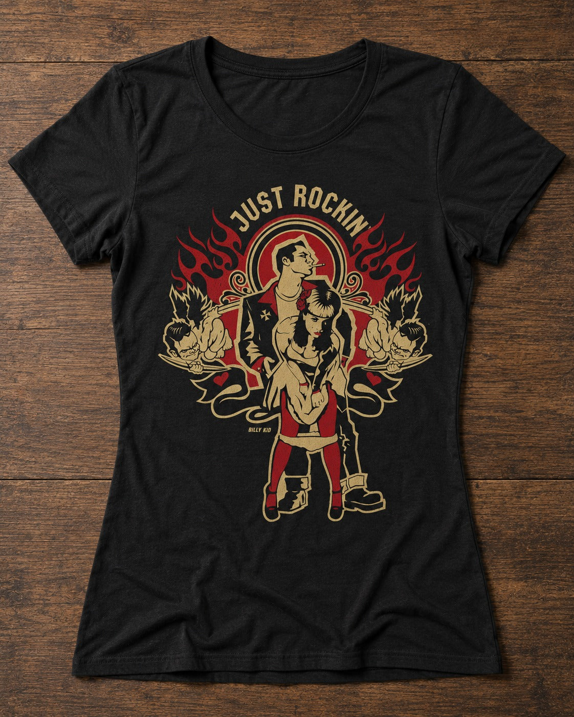 A black short-sleeve graphic t-shirt laid flat on a dark wood background. The shirt features a vintage-style tattoo-style graphic of a woman and man with flames, roses, and the text 'JUST ROCKIN' in gold and red.