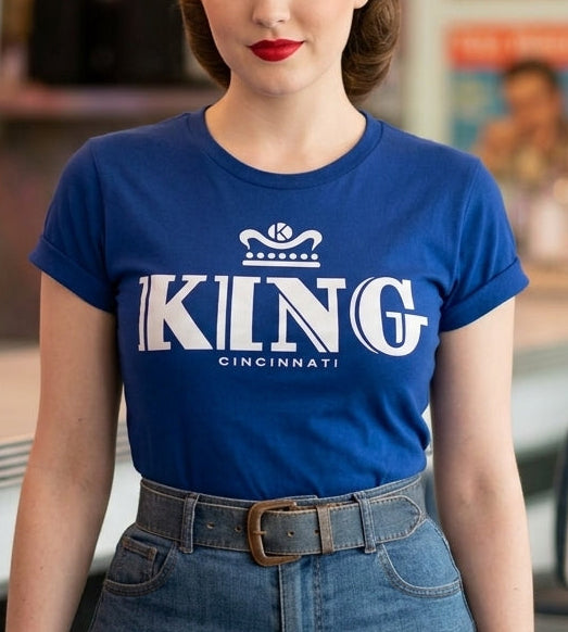 A woman with vintage-style brown hair stands in a retro diner wearing a blue short-sleeve graphic T-shirt with 'KING RECORDS CINCINNATI printed in sinver across the chest, paired with blue jeans and a brown belt.