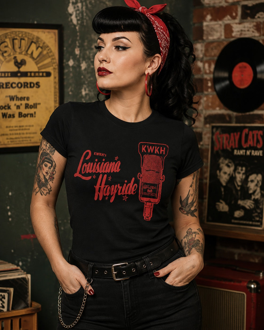A woman with vintage-style black hair, a red bandana, and tattoos wears a black graphic T-shirt featuring red text reading 'Louisiana Hayride' and a red KWKH microphone graphic. She stands in a retro-themed room with posters, a vinyl record, and an amplifier in the background.