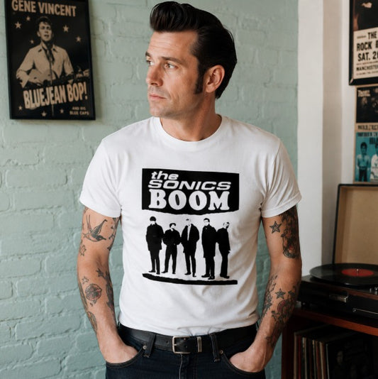 A man wearing a white graphic t-shirt with a black print of 'The Sonics Boom' and the band members, standing in front of a light blue brick wall with vintage posters.