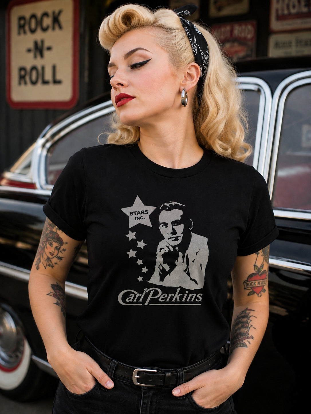 A woman with vintage-style blonde hair and tattoos wears a black graphic T-shirt featuring a white print of Carl Perkins and the text 'Stars & Stripes' and 'Carl Perkins.' She is posed in front of a black car with a rustic sign in the background.