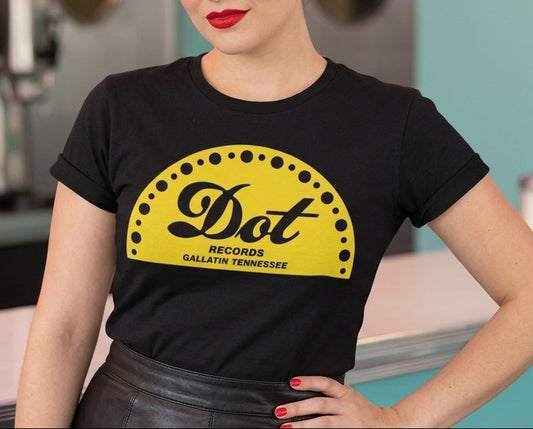 A woman with vintage-style curled hair stands in a retro diner setting wearing a black t-shirt with a yellow 'Dot Records' graphic on the front.