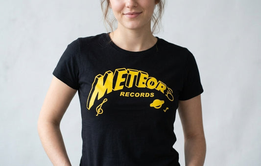 A woman wearing a black short-sleeve t-shirt with a yellow 'Meteors Records' graphic on the front, set against a light gray textured wall.