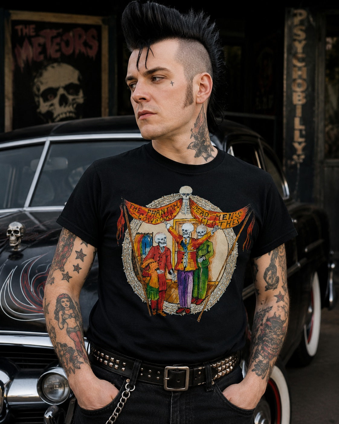 A man with a black mohawk and tattoos wears a black graphic t-shirt featuring the band 'Cult Leader' with skeleton figures. He is posed in front of a vintage car with rock-and-roll themed posters in the background.