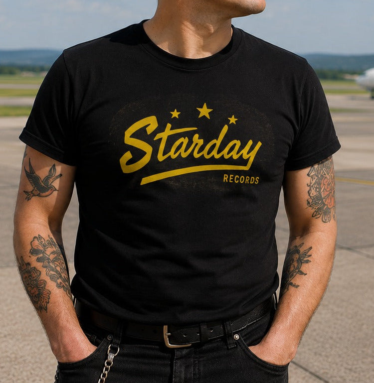 A man wearing a black graphic t-shirt with a yellow 'Starday Records' logo stands on an airport tarmac with an airplane and cloudy sky in the background.