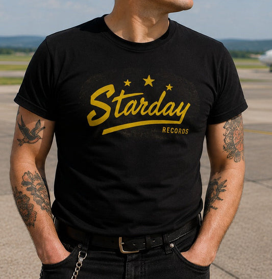 A man wearing a black graphic t-shirt with a yellow 'Starday Records' logo stands on an airport tarmac with an airplane and cloudy sky in the background.
