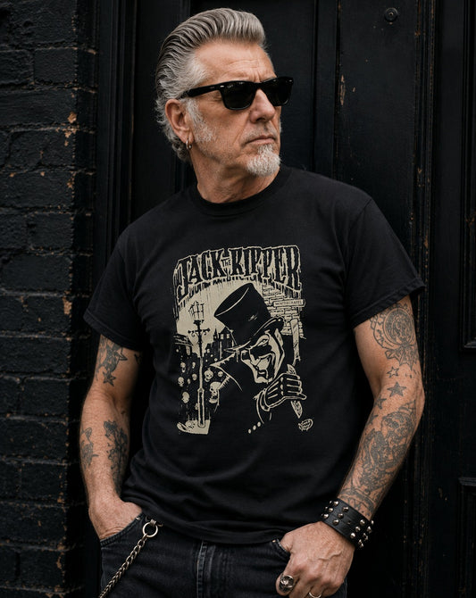 A man with gray hair, sunglasses, and tattoos leans against a black wooden door and brick wall, wearing a black graphic t-shirt with a beige 'Jack the Ripper' illustration.